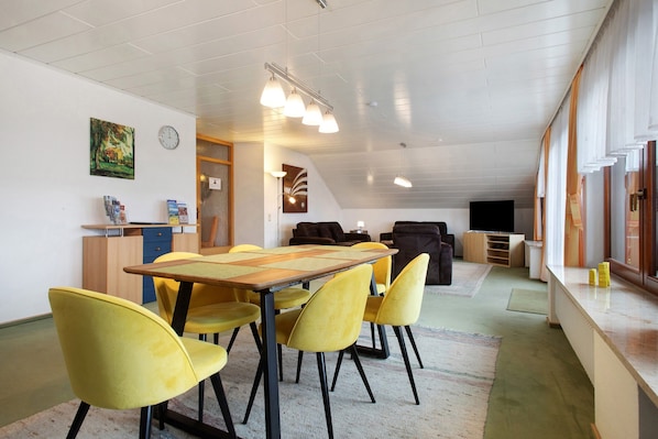 Un comedor con mesa de madera y sillas amarillas, una zona de cocina con armarios y microondas, una sala de estar con sofá y televisor, y un cuadro en la pared.
