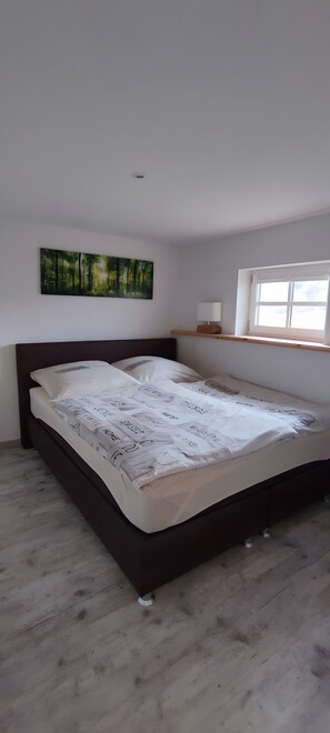 Un dormitorio con una cama, cabecera, un cuadro en la pared, una mesita de noche y una ventana.