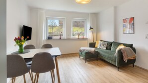 Ein modernes Wohnzimmer mit Esstisch, Stühlen, einer Couch, einem Fernseher und einem Fenster mit Blick auf einen Park.