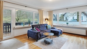 A modern living room with a sectional sofa, a round coffee table, and large windows with curtains. - Apartment 'Stötenberg' with Mountain View, Private Garden and Wi-Fi (Schmallenberg)