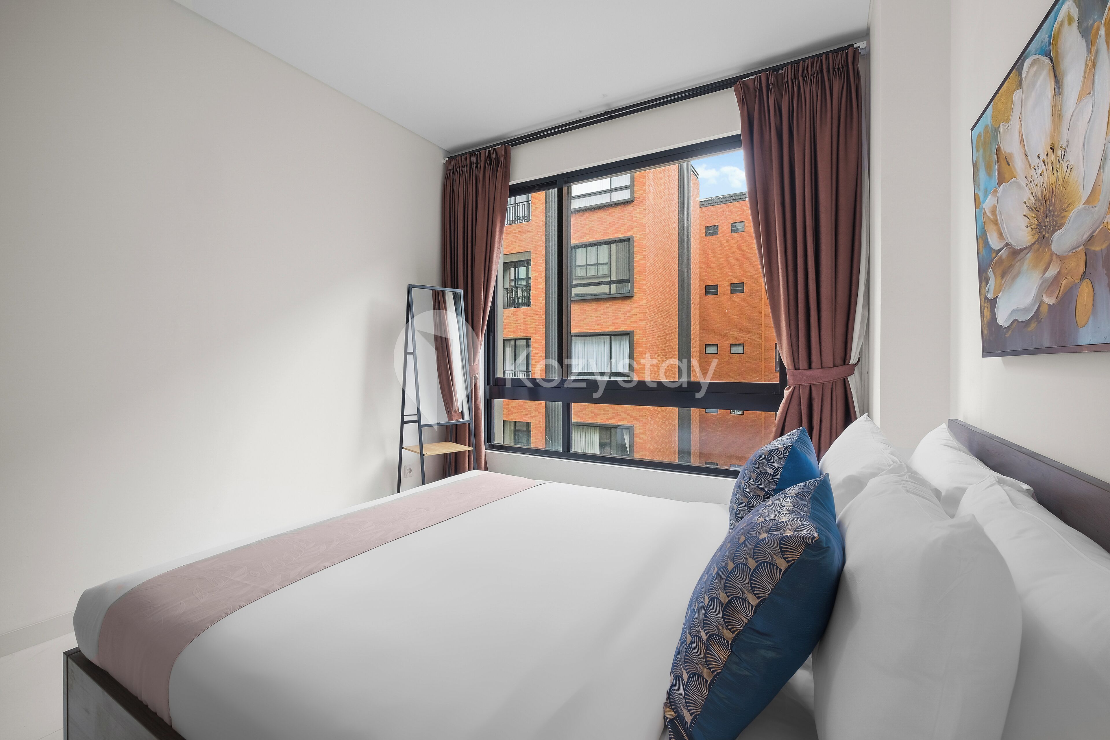 A hotel room with a large bed, a window with curtains, a ladder, and a painting on the wall.