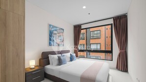 A modern bedroom with a large bed, a nightstand, a wall-mounted painting, and a view of a brick building through a window with curtains.