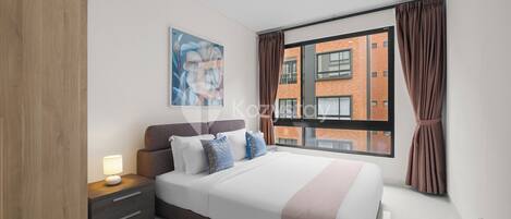 A modern bedroom with a large bed, a nightstand, a wall-mounted painting, and a view of a brick building through a window with curtains.