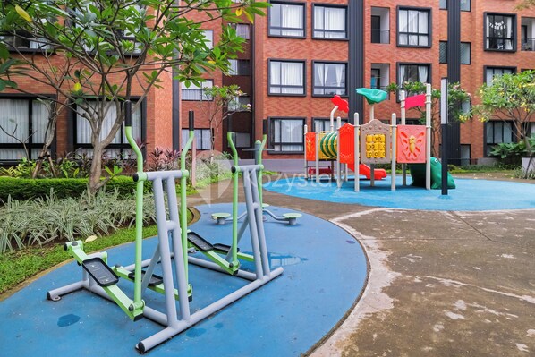 Un área de gimnasio al aire libre con equipo de ejercicio, una estructura de juegos infantiles y un edificio con varias ventanas al fondo.