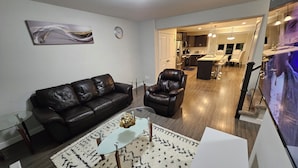 Ein modernes Wohnzimmer mit einer schwarzen Ledersofa, einem Ledersessel, einem Glastisch und einem gemusterten Teppich. Ein Gemälde hängt an der Wand, und im Hintergrund ist eine Küche zu sehen.