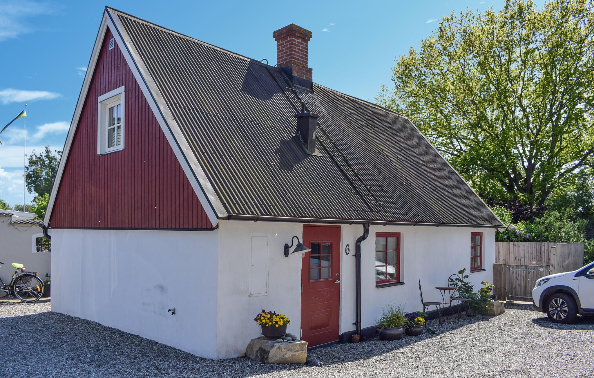 Een klein, gelijkvloers huis met een rode en witte gevel, een schuin dak en een schoorsteen. Voor het huis staan potplanten en links staat een fiets geparkeerd.