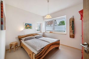 A bedroom with a wooden bed, a small stool, a painting, and a window with a view.