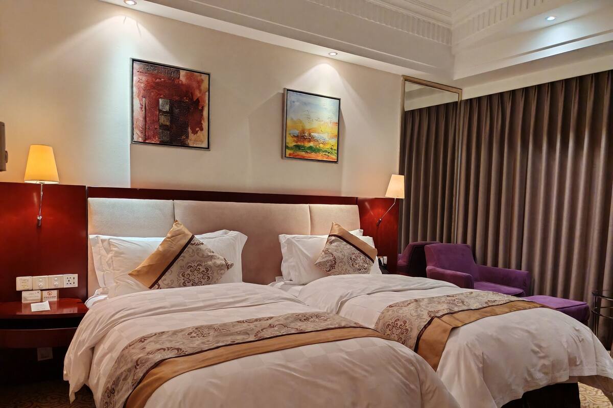 Quarto de hotel com duas camas, mesa de cabeceira com abajur, espelho e vista para o exterior através de cortinas.