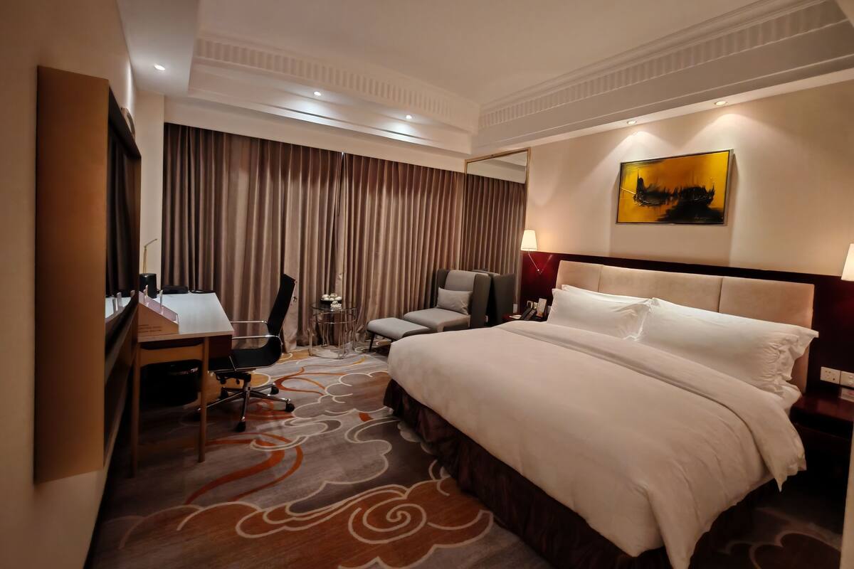 Quarto de hotel com uma cama grande, uma escrivaninha com cadeira, uma mesinha e outra cadeira.