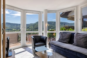 A balcony with a view of green hills, a black leather chair, a blue sofa, and a glass coffee table.