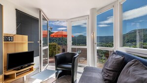 A modern living room with a flat-screen TV, a black leather chair, a sofa, and a balcony with an orange umbrella. - Apartment 'Fewo2' with Mountain View, Private Terrace and Wi-Fi (Bad Urach)