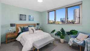 A bedroom with a bed, bedside tables, a bench, a chair, and two potted plants. - Ambassador Beachfront Apartments (Burleigh Heads)