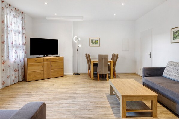 A modern living room with a flat-screen TV, wooden furniture, a dining table with chairs, a sofa, and a floor lamp.