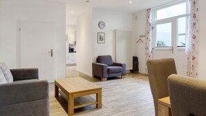 En moderne stue med sofa, lænestol og sofabord. I baggrunden ses et køkken med et ur og et billede i ramme på væggen.
