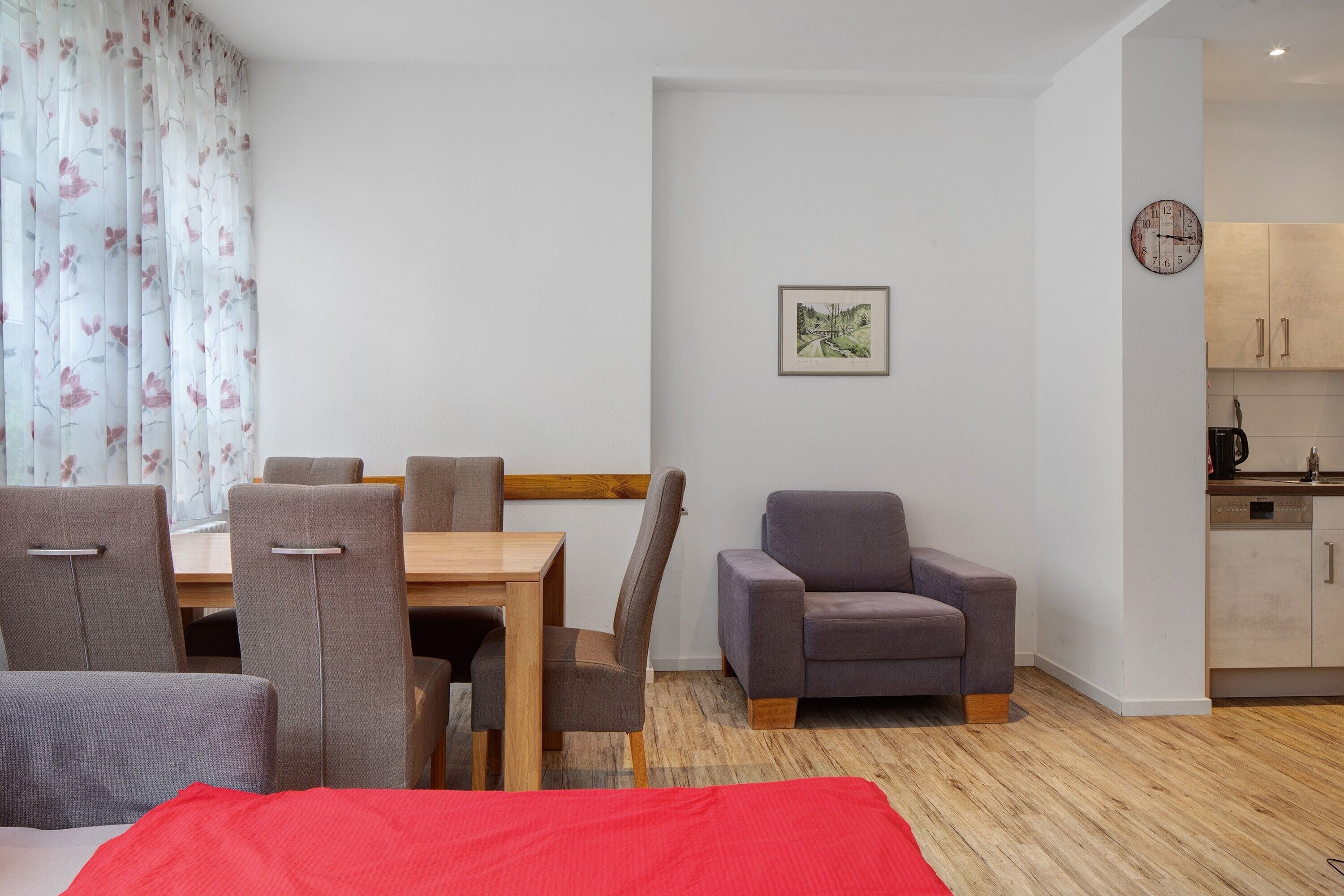 Une chambre moderne avec une table à manger, des chaises, un canapé, un lit recouvert d’une couverture rouge, une horloge et un tableau encadré au mur.