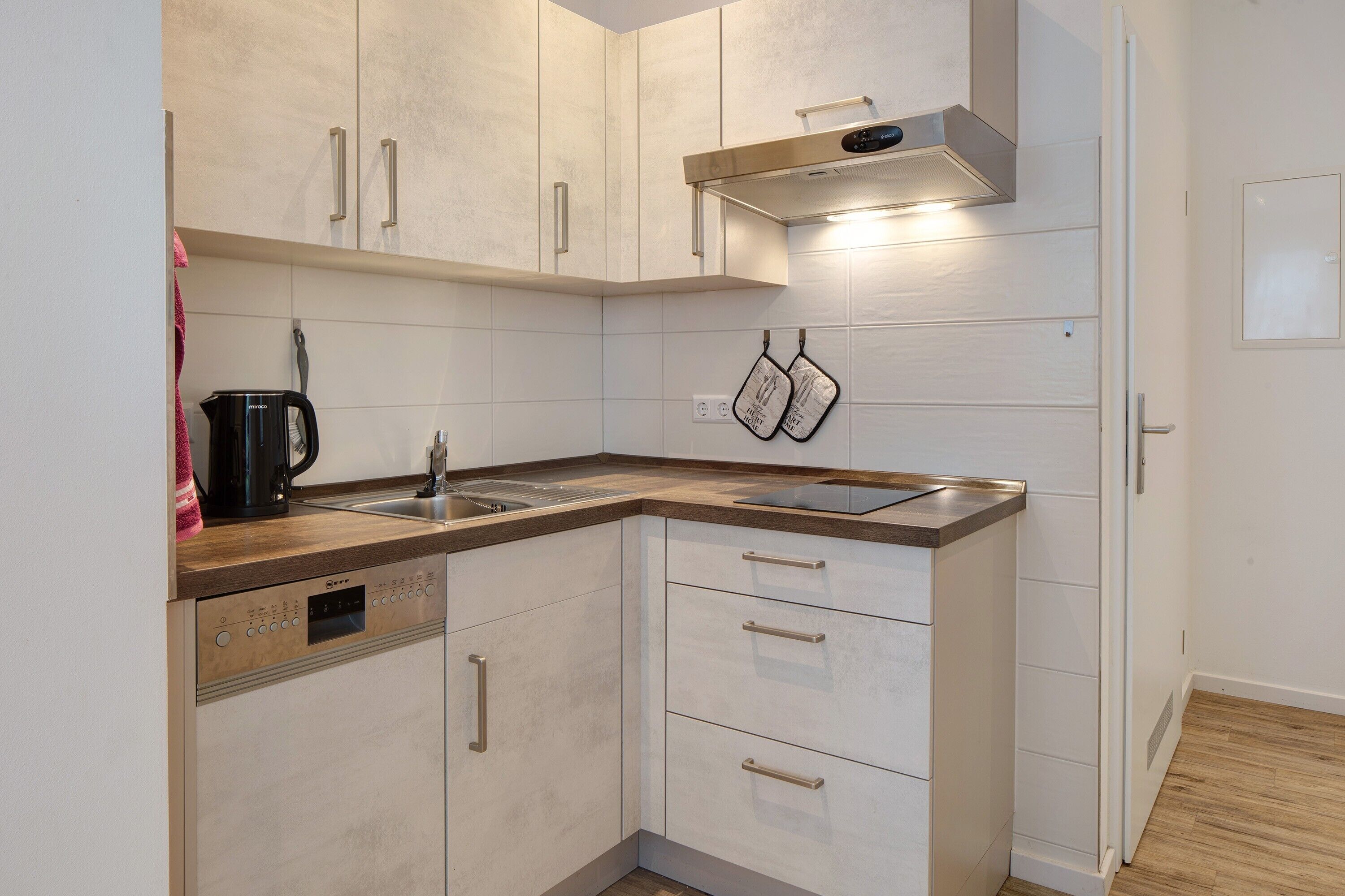 Une cuisine compacte avec des meubles blancs, un lave-vaisselle, une hotte de cuisinière en acier inoxydable et un plan de travail en bois.