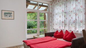 Een slaapkamer met een groot raam, een bed met rode beddengoed, een stoel en een kleine tafel.