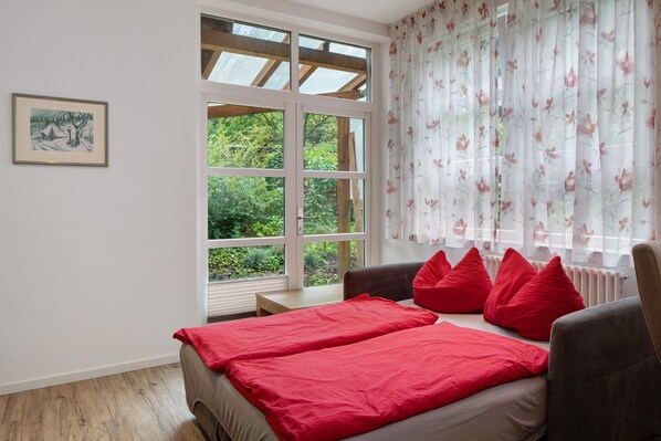 A bedroom with a large window, a bed with red bedding, a chair, and a small table.
