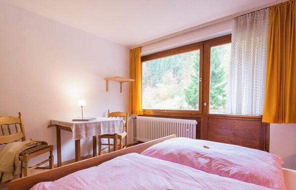 Ein Schlafzimmer mit zwei Betten, einem Holzstuhl, einem kleinen Tisch mit Lampe und einem Fenster mit Vorhängen.