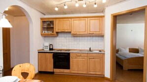 A compact kitchen with wooden cabinets, a stove, and a sink. There is a dining table with chairs and a bed in the adjacent room. - Apartment 'Ferienwohnung Schwalbennest' with Mountain View, Balcony and Wi-Fi (Mühlenbach)