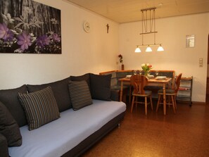 A cozy living room with a sofa, a dining table with chairs, a clock, and a cross on the wall.