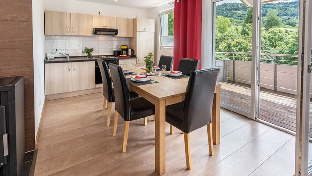 A modern kitchen with wooden cabinets, a dining table with chairs, and a balcony with a red curtain. - Apartment 'Teufelsmühlblick' with Mountain View, Balcony and Wi-Fi (Loffenau)