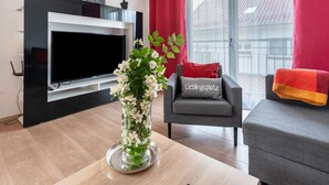 A modern living room with a flat-screen TV, a grey sofa with a red cushion, a small table with a vase of flowers, and a window with red curtains. - Apartment 'Teufelsmühlblick' with Mountain View, Balcony and Wi-Fi (Loffenau)