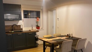 Une cuisine moderne avec des meubles de rangement sombres, une table à manger en bois et une fenêtre équipée de stores.