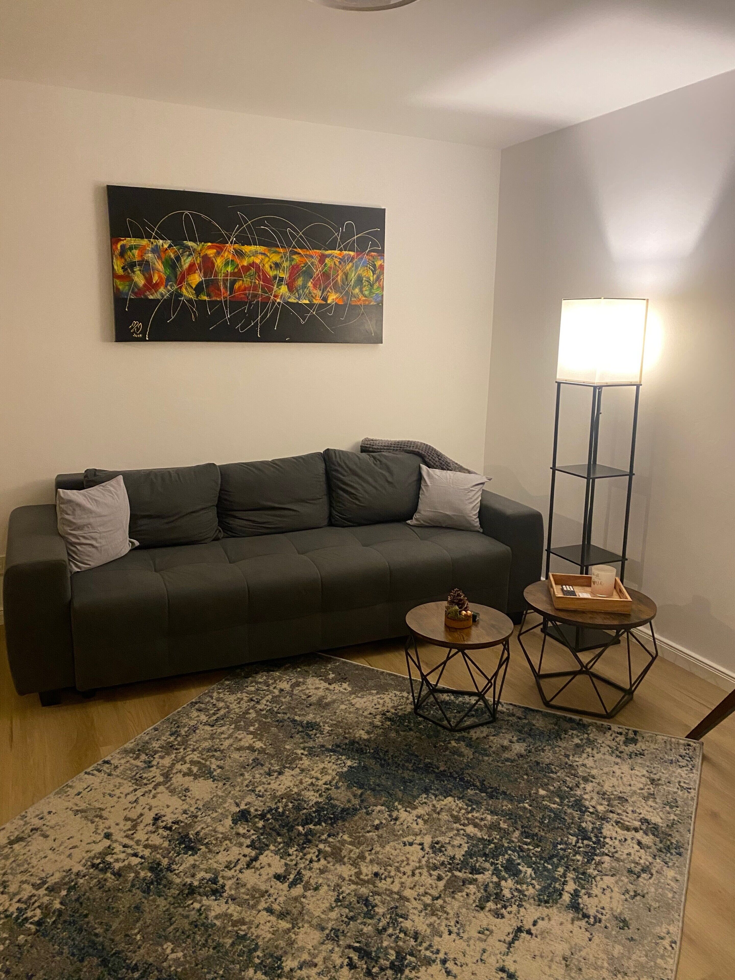 Un salon avec un canapé gris foncé, un tableau abstrait aux couleurs vives, un grand lampadaire et une table basse ronde sur laquelle se trouve un plateau.