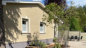 Un bâtiment à l’extérieur de couleur beige, doté de deux fenêtres, d’une terrasse pavée et d’un petit jardin.