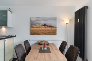 Een eetgedeelte met een houten tafel, vier stoelen, een vaas met bloemen en een schilderij van een strand.