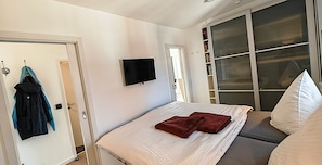 Quarto com cama, televisão montada na parede, guarda-roupa e porta de correr de vidro.
