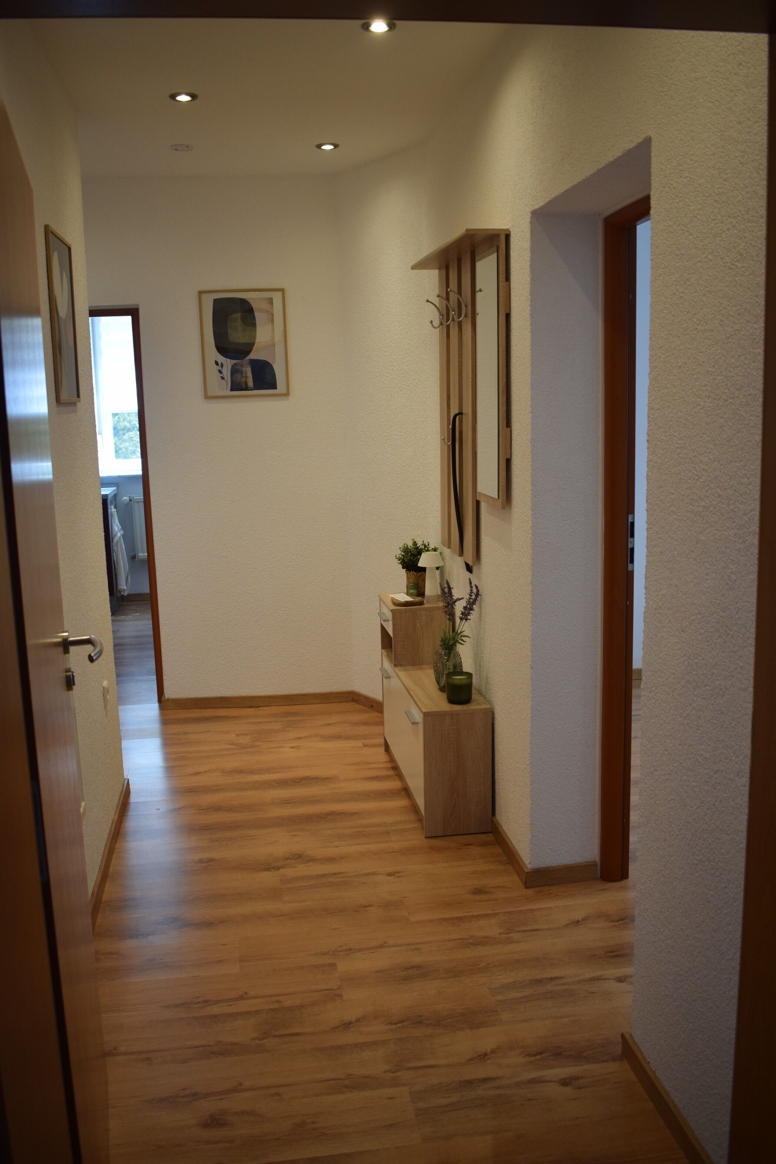 Ein Flur mit Holzboden, einer Tür links, einer kleinen Pflanze auf einem Holzständer und einem gerahmten Bild an der Wand.