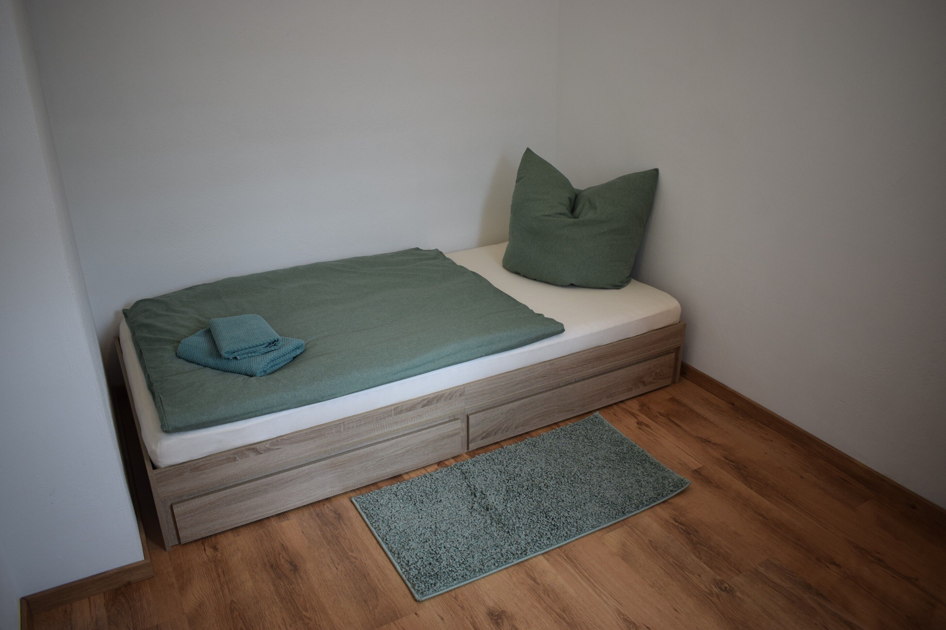 A small bed with a green blanket and pillow, a wooden drawer, and a grey rug on a wooden floor.