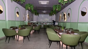 Un restaurante con sillas tapizadas de verde, mesas dispuestas con copas de vino y platos, y una persona al fondo.