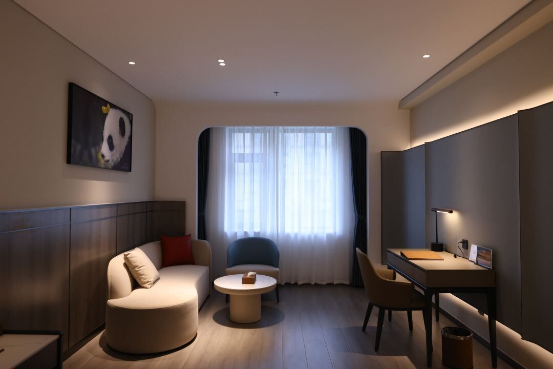 A modern hotel room with a sofa, armchair, round coffee table, and a desk with a lamp.