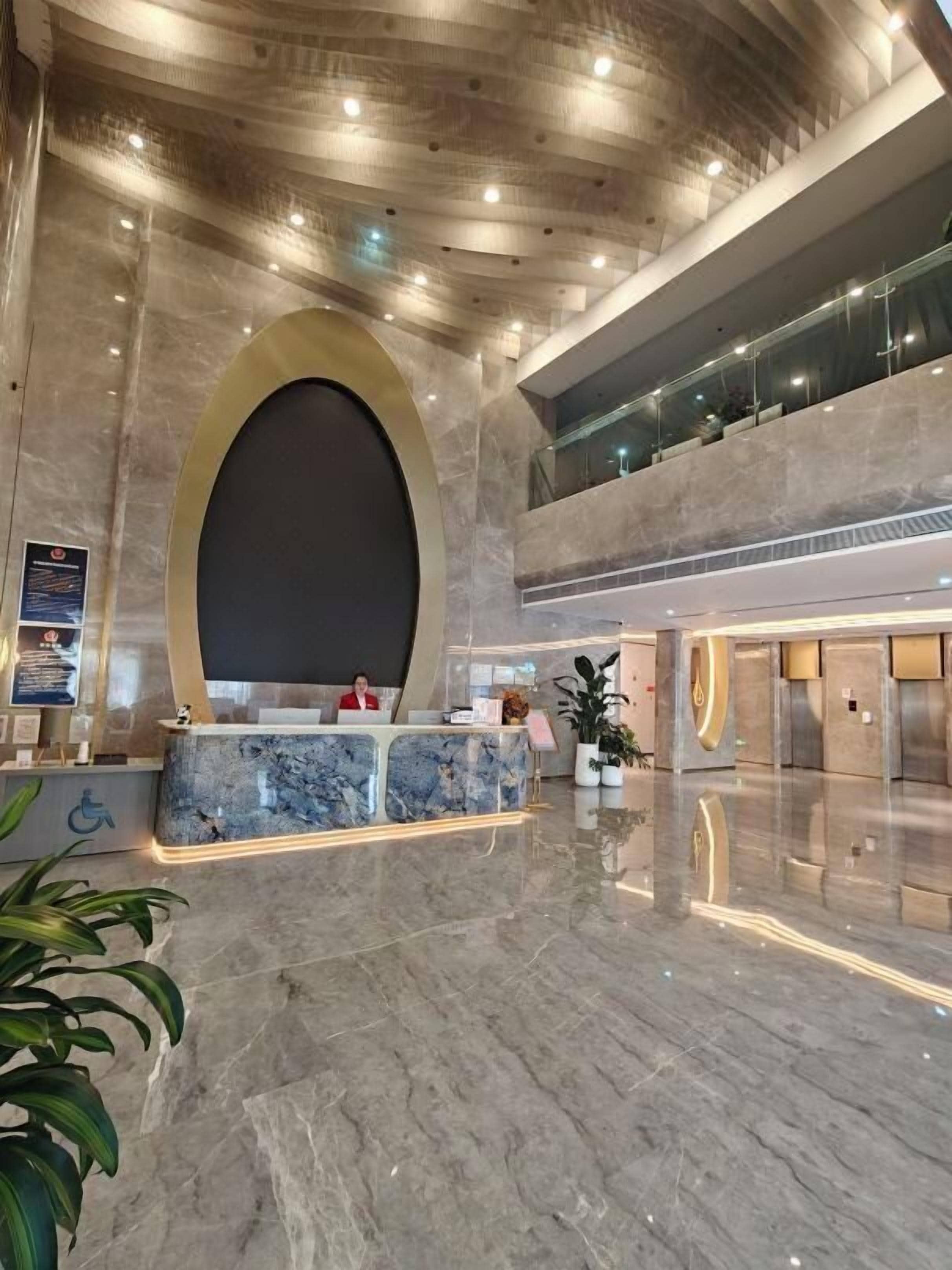 Una hall d'albergo moderna con una reception in marmo, un grande specchio ovale e una ringhiera in vetro che si affaccia su un livello inferiore.