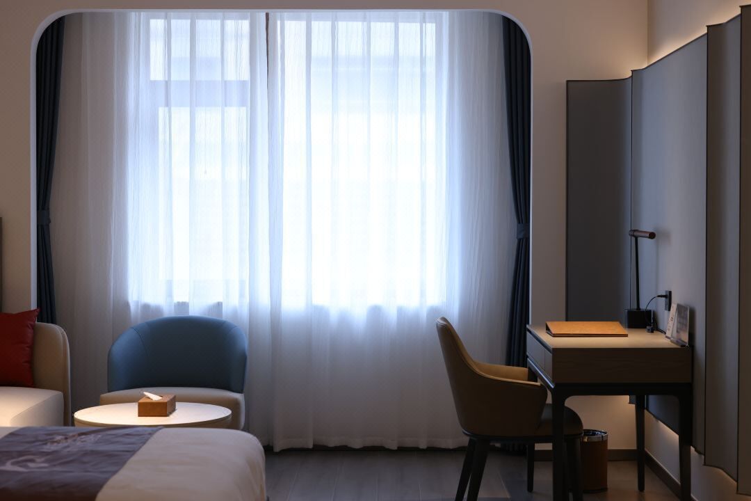 A hotel room with a bed, two chairs, a desk, and a large window with sheer curtains.