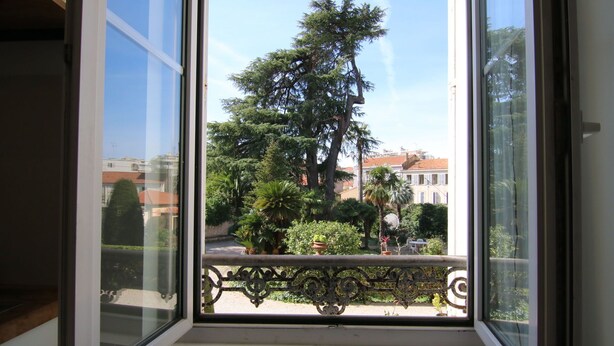 Une fenêtre donnant sur un balcon, un grand arbre et des bâtiments en arrière-plan.