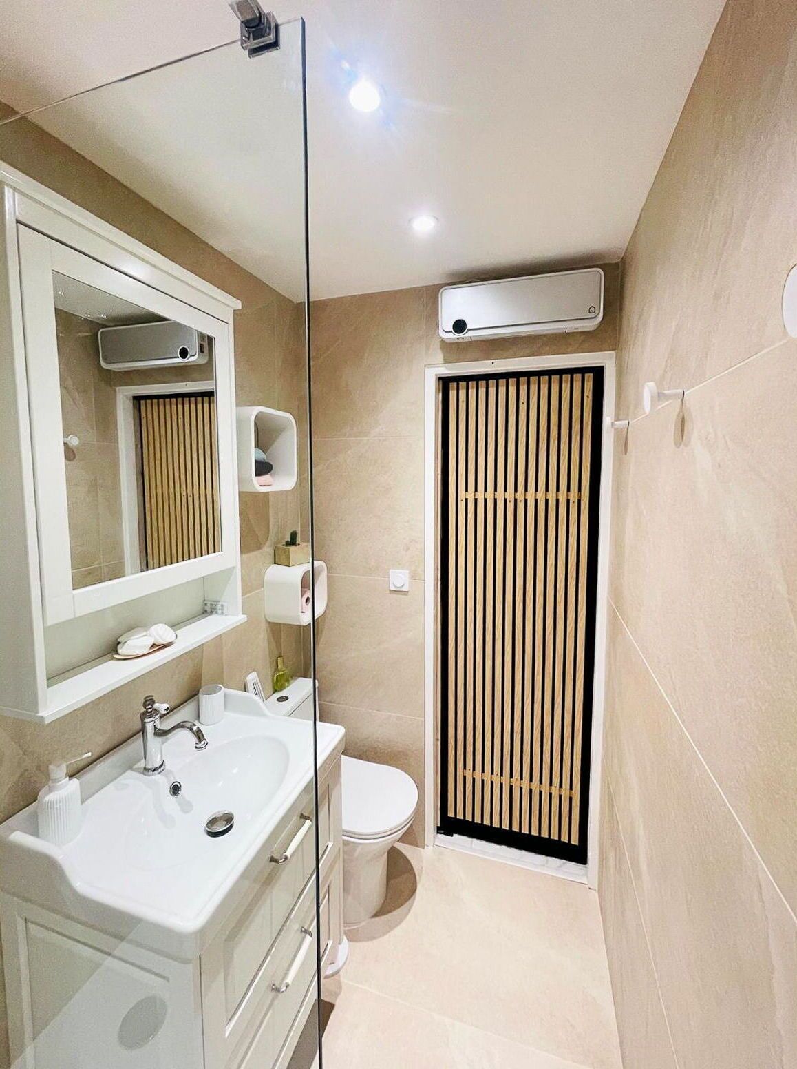 Un baño moderno con lavamanos, espejo y retrete blancos. Se observan un dispensador de toallas y una unidad de ventilación en la pared.