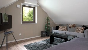 A modern living room with a flat-screen TV on a stand, a glass coffee table, a grey sofa with patterned cushions, a large window with a view of greenery, and an air conditioning unit on the wall.