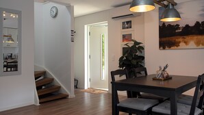 A dining area with a wooden table, chairs, and pendant lights. A staircase leads to another room, and a clock is visible on the wall.