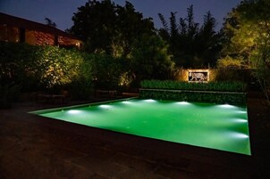 Piscina com borda iluminada em verde, rodeada por vegetação exuberante e um muro de pedra.