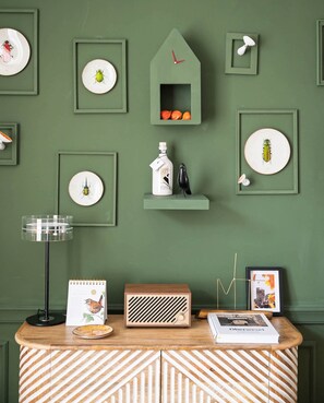 A room with a green wall, framed insect illustrations, a small shelf with a bottle and a bird figurine, a wooden radio, and a framed book on a wooden table. - Ferien Apartment Schinderhannes (Oberwesel)