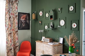 Une pièce avec un mur vert, un tableau représentant un renard encadré, une chaise rouge, un meuble contenant des objets décoratifs et un vase vert avec des fleurs séchées.