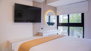 Un dormitorio moderno con una cama grande, un televisor montado, una mesita de noche y una ventana con vistas a edificios y árboles.