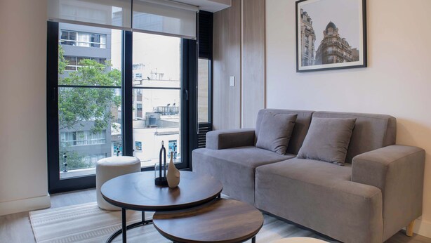 Una sala de estar moderna con un sofá gris, una mesa redonda de madera y un ventanal que ofrece vistas a edificios y árboles.
