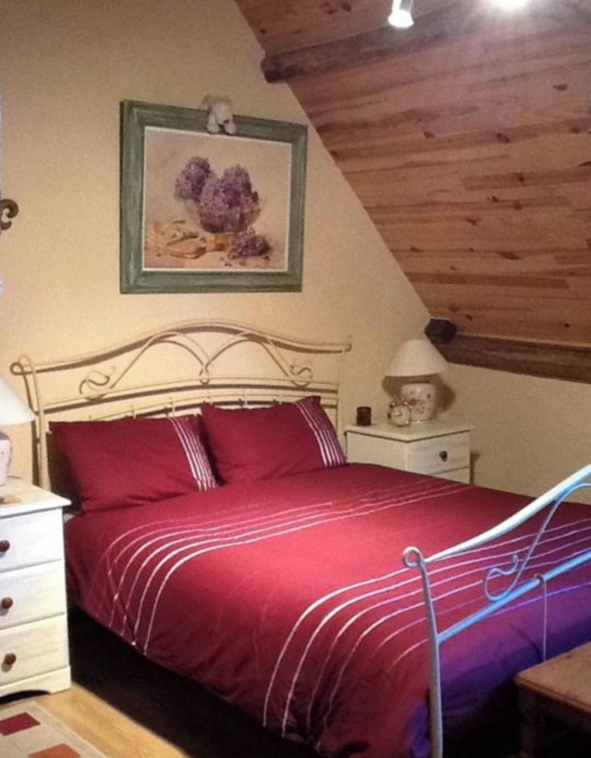 A bedroom with a metal bed frame, a red bedspread, a nightstand with a lamp, and a framed picture on the wall.