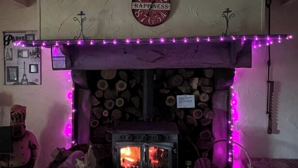 Ein gemütlicher Raum mit Kaminofen und einem Schild mit der Aufschrift 'HAPPINESS', beleuchtet von violetten Lichterketten.