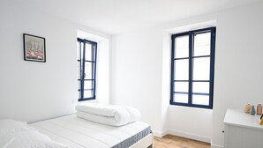 Un dormitorio moderno con una cama, dos ventanas, un tocador blanco y un cuadro colgado en la pared.